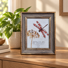 Load image into Gallery viewer, A framed botanical art print featuring a red dragonfly and dandelion on a light background, styled on a wooden table next to a potted plant.