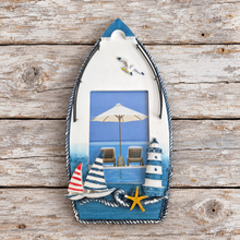Load image into Gallery viewer, A nautical-themed wooden boat wall decor piece featuring a blue and white beach scene with a lighthouse, sailboat, umbrella, seagull, and starfish, displayed on a rustic wooden plank background.