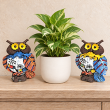 Load image into Gallery viewer, Two decorative owl figurines with colorful patterned scarves sit on either side of a potted green plant on a light wood surface against a beige wall.