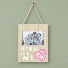Load image into Gallery viewer, A light wood hanging plaque with a rope hanger, featuring a photo slot with a family picture and the text 'TEACHING is a WORK' with a pink heart accent, set against a sage green wall.