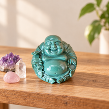 Load image into Gallery viewer, A glossy turquoise Laughing Buddha figurine sits on a wooden table, styled with crystal points and a purple amethyst cluster. A blurred green plant is visible in the background.