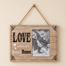 Load image into Gallery viewer, A rustic wooden hanging wall sign with a photo slot, featuring the text 'LOVE makes our house a Home' and a black-and-white family photo, hanging from a rope against a beige wall.