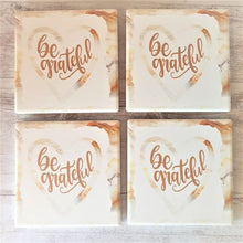 Load image into Gallery viewer, Add a touch of positivity to your home decor with our Be Grateful Coasters. This set of 4 table coasters serves as a reminder to practice gratitude every day. Perfect for entertaining, or as a thoughtful gift for loved ones. Gratitude is always in style!  Ceramic | 10 x 10 cm | Cork non slip backing | White gift box with lid | Set of 4 same design | Square.